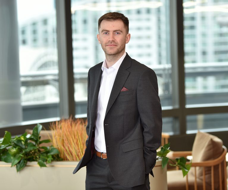 A man in a dark grey suit with a white shirt, standing in a modern office space with large windows, plants, and cityscape in the background.