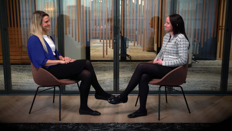 Two women sitting facing each other in brown chairs with glass doors behind them, appearing to have a conversation in a modern indoor setting.
