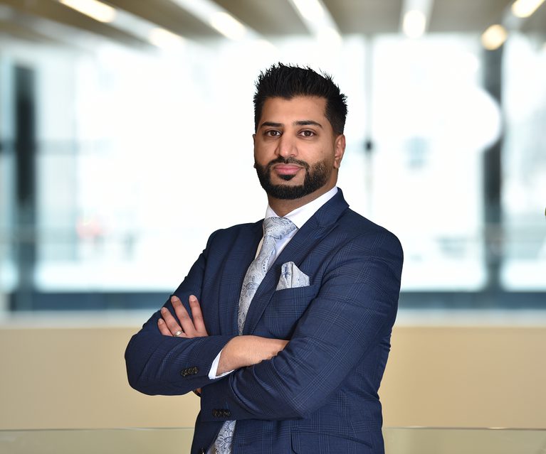 A confident man with a beard and styled hair, wearing a navy suit with a patterned tie and pocket square, stands with arms crossed in a modern office space.