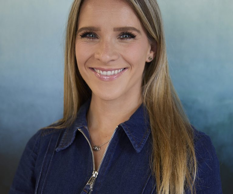 Portrait of a person with long blonde hair, wearing a blue denim jacket, smiling at the camera against a blue gradient backdrop.