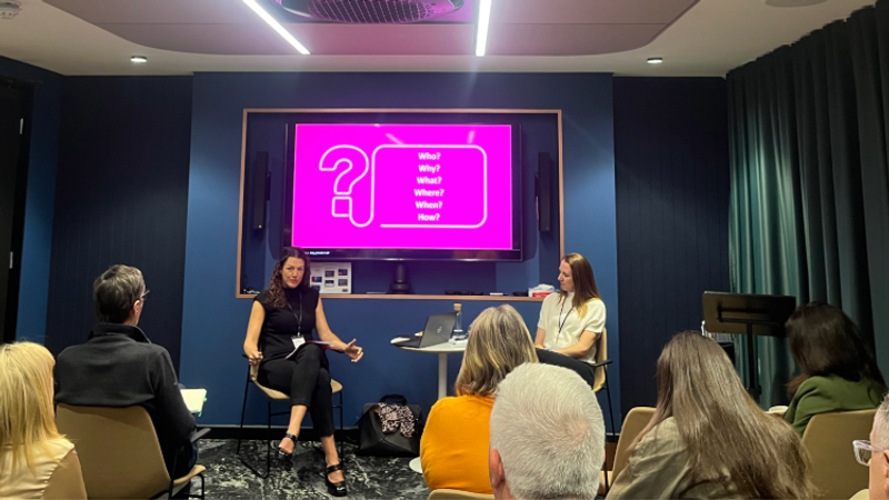 Two speakers on a small stage in a conference room, one talking with a laptop nearby; a bright pink screen behind shows a large question mark and Who? Why? What? Where? When? How?