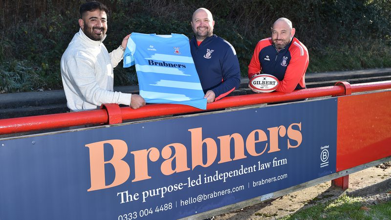 Three men are standing behind a red fence, smiling. One holds a blue rugby jersey with "Brabners" on the front, and another is holding a rugby ball.