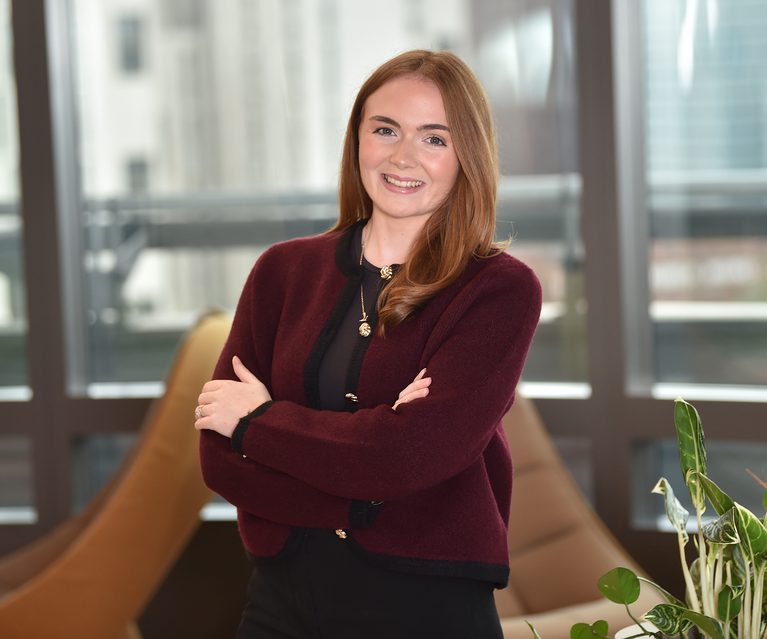 A woman with long, wavy auburn hair smiling with arms crossed, wearing a burgundy and black cardigan, in a modern office with large windows and a green plant.