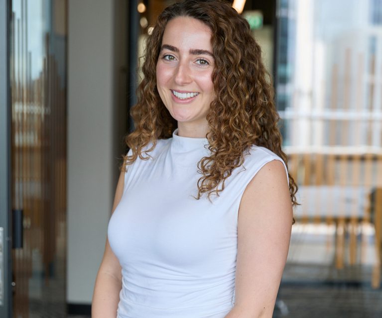 Person with curly brown hair, smiling, wearing a white sleeveless top and dark trousers, standing in a bright, modern indoor space with glass doors.