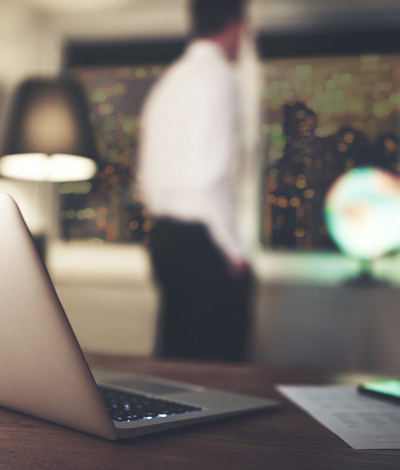 A laptop on a wooden desk with a blurred person standing in front of a window displaying city lights, and a globe and mouse on the desk.