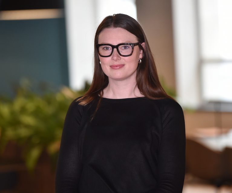 A woman with long brown hair, wearing black glasses and a black top, standing indoors with large windows and blurred greenery in the background.