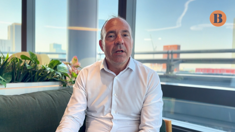 A person with short grey hair in a white shirt sits on a dark sofa in a bright office, with plants on the left and a cityscape visible through large windows; an orange circle with a blue B sits top-right.