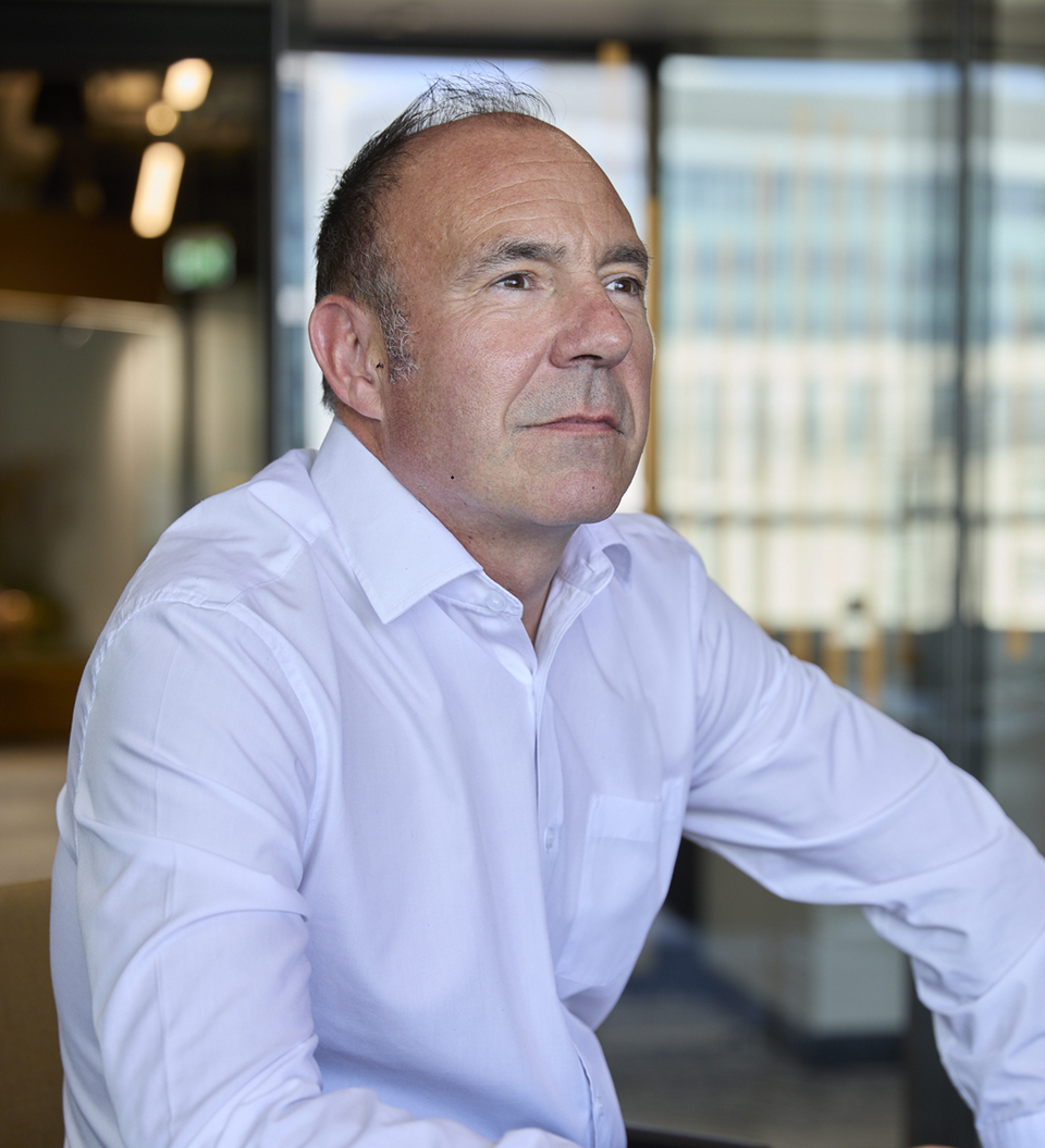A middle-aged person wearing a white shirt sits at a dark desk in a modern office, gazing thoughtfully to the left with glass walls and distant buildings in view.