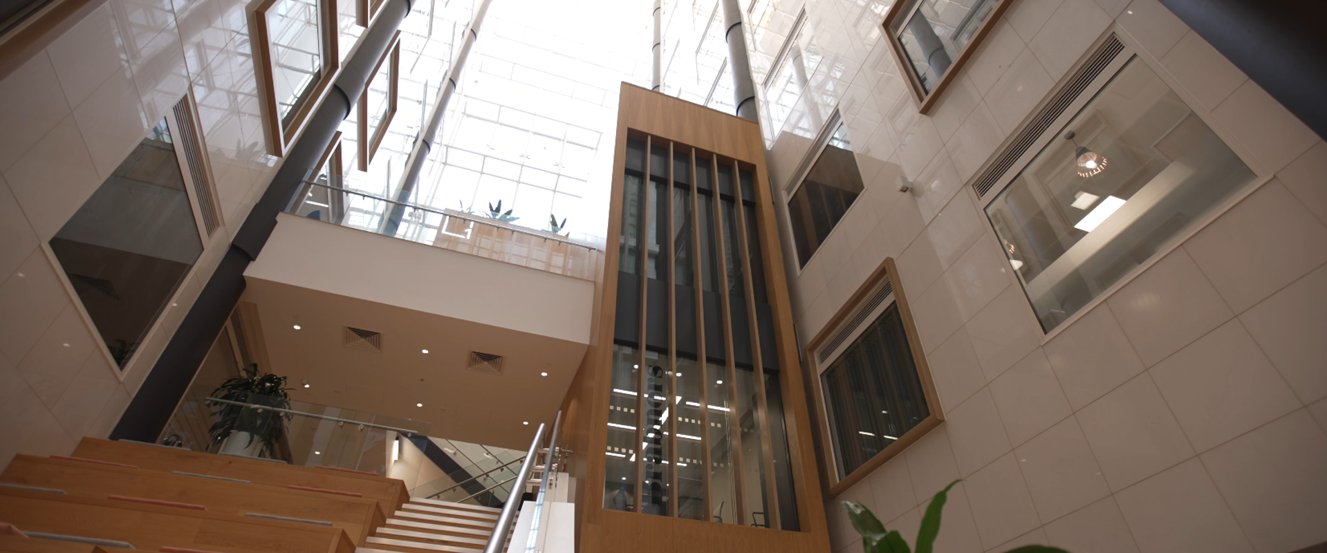 A modern indoor atrium with a glass ceiling, featuring wooden staircase, framed windows on walls, and a central glass-enclosed elevator shaft.