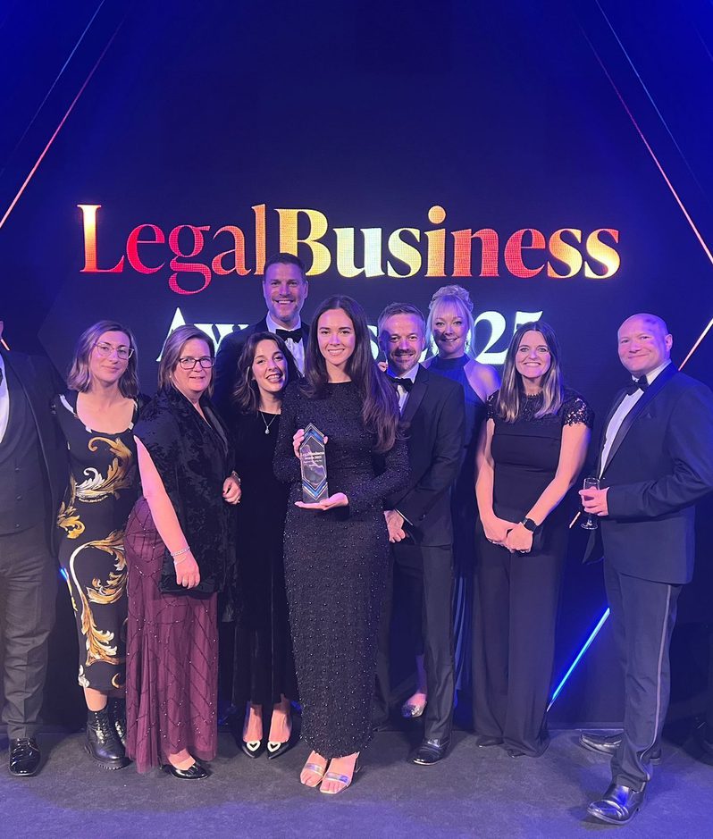 Group of nine people in formal attire posing with an award on stage at the Legal Business Awards 2022 event.