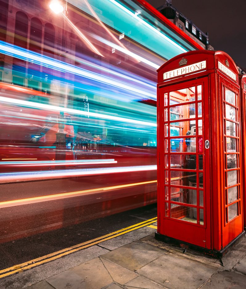 A red traditional British telephone box on a city street at night, with colourful light streaks from passing vehicles and buildings illuminated in the background.