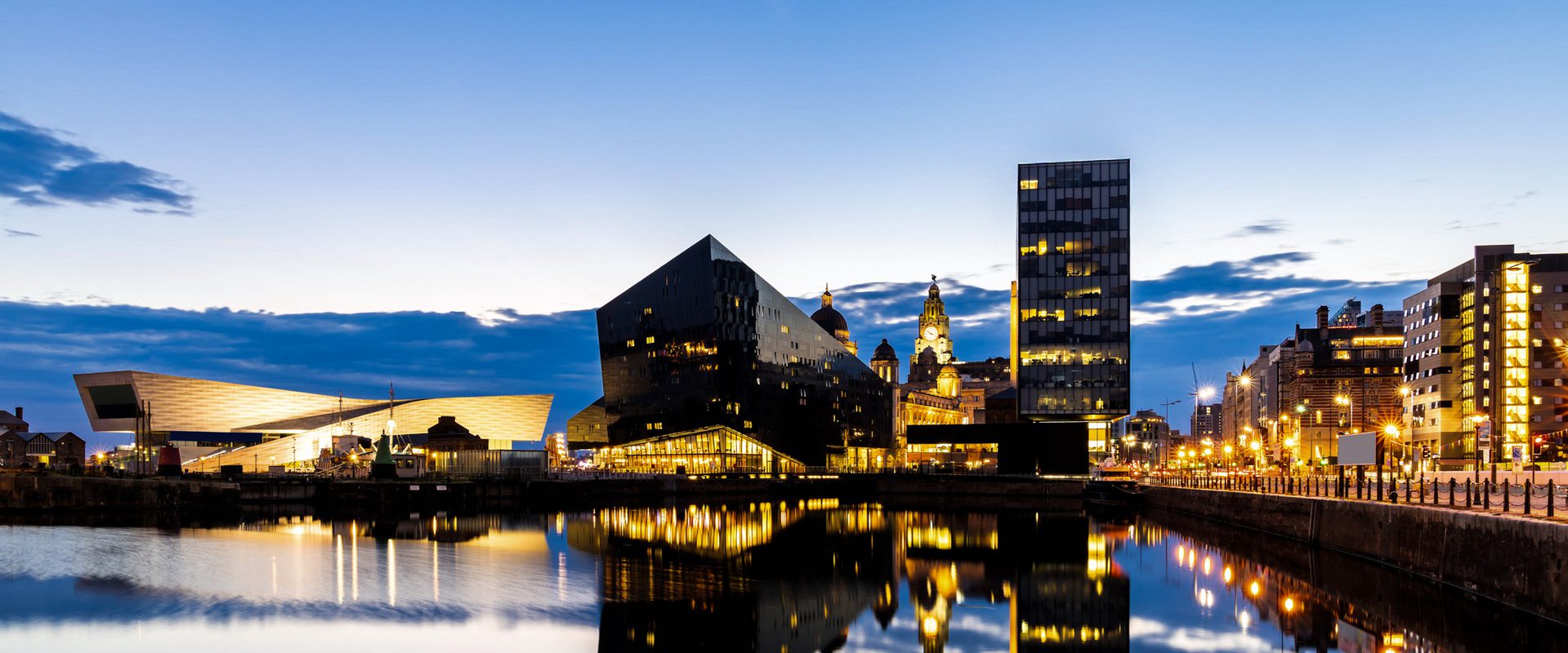 City skyline at dusk with modern buildings reflected in a calm waterfront, illuminated by streetlights and interior lights against a blue sky.