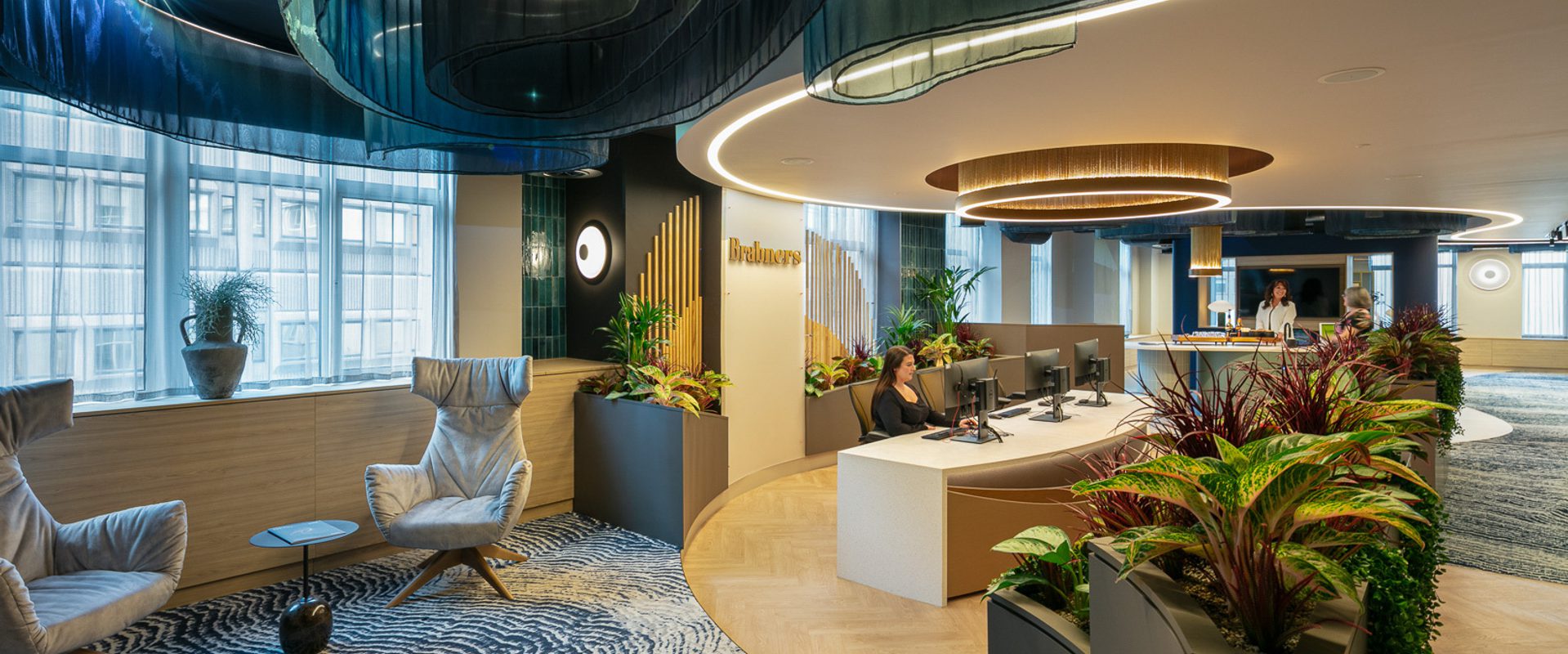 Modern office reception with curved illuminated ceiling elements, circular light rings, lush planters, and a curved desk where staff assist visitors.