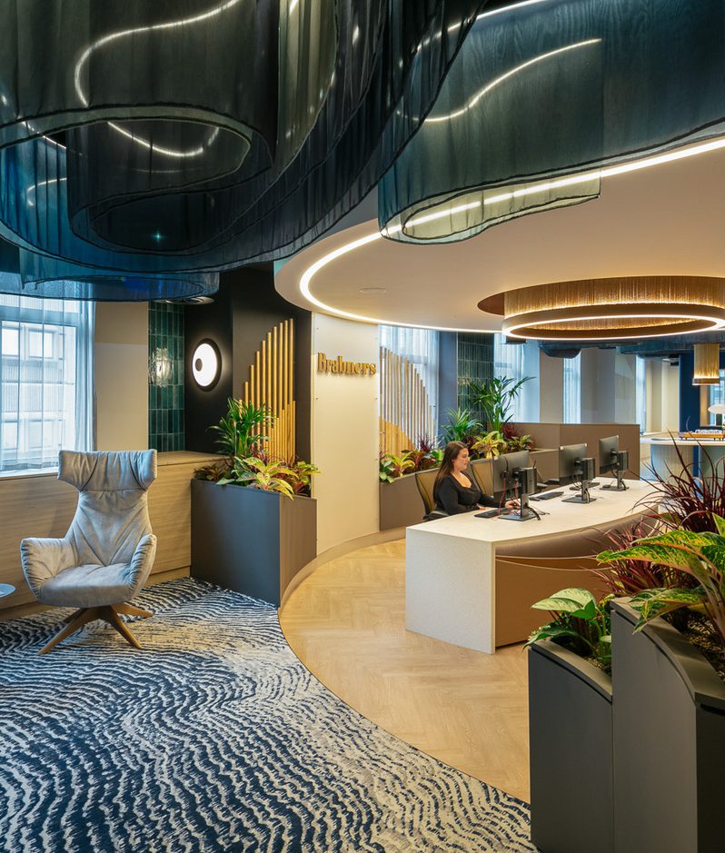 Modern office reception with curved illuminated ceiling elements, circular light rings, lush planters, and a curved desk where staff assist visitors.