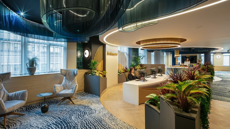 Modern office reception with curved illuminated ceiling elements, circular light rings, lush planters, and a curved desk where staff assist visitors.