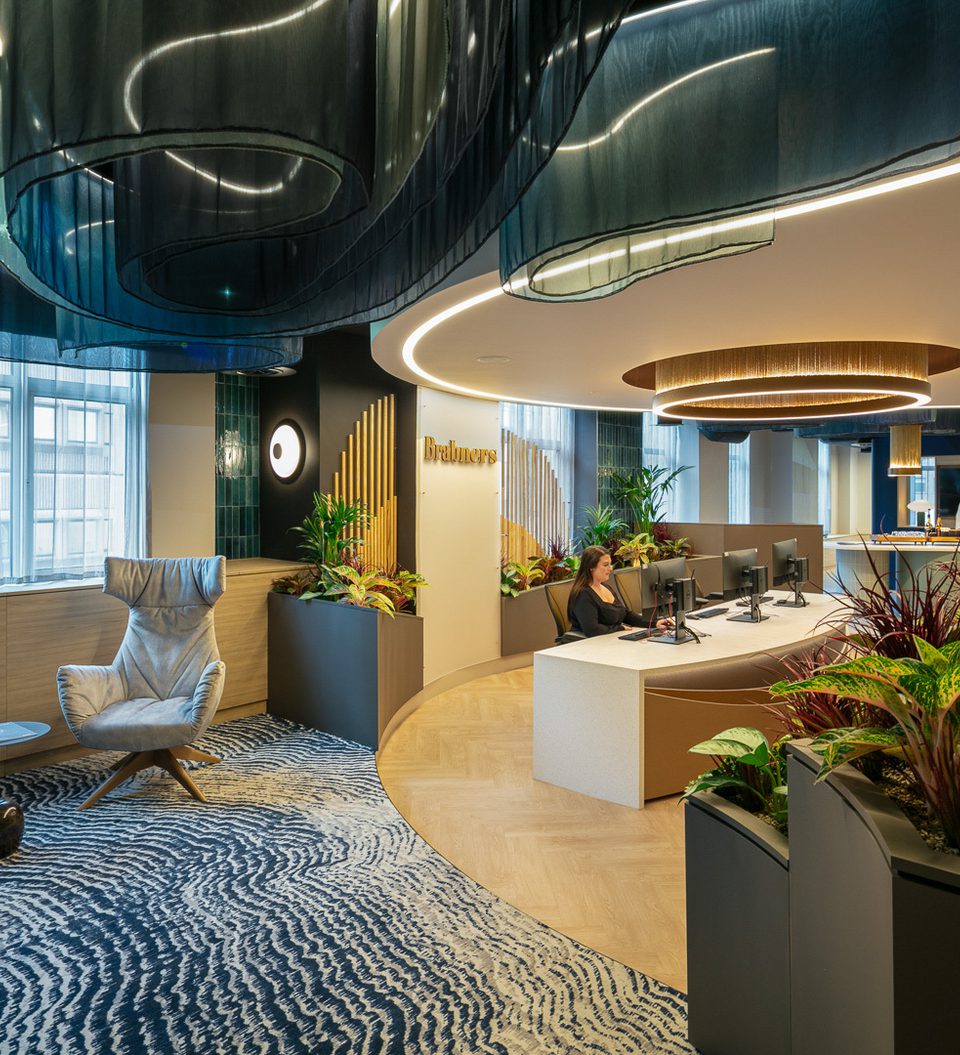 Modern office reception with curved illuminated ceiling elements, circular light rings, lush planters, and a curved desk where staff assist visitors.