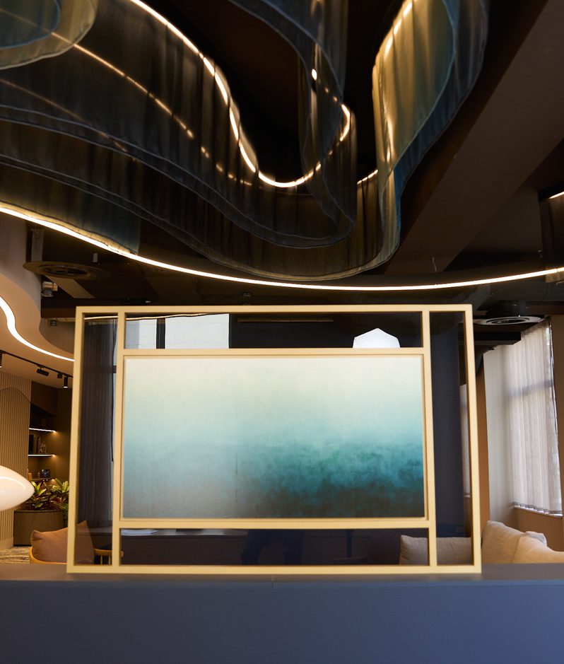 Modern interior lounge, wavy ceiling with backlit lines, gold-framed gradient screen, blue curved seating, and a white mushroom lamp by windows.
