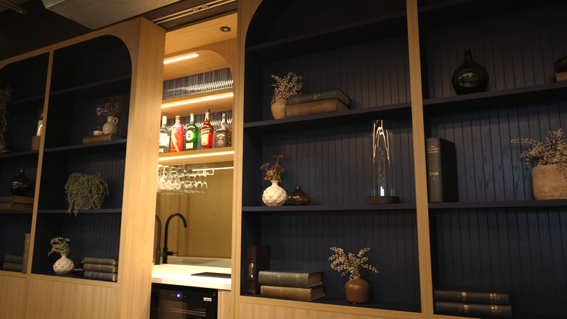 A wooden and dark blue shelving unit with books, vases, and decorative items, and a small kitchen area with a sink and hanging wine glasses.