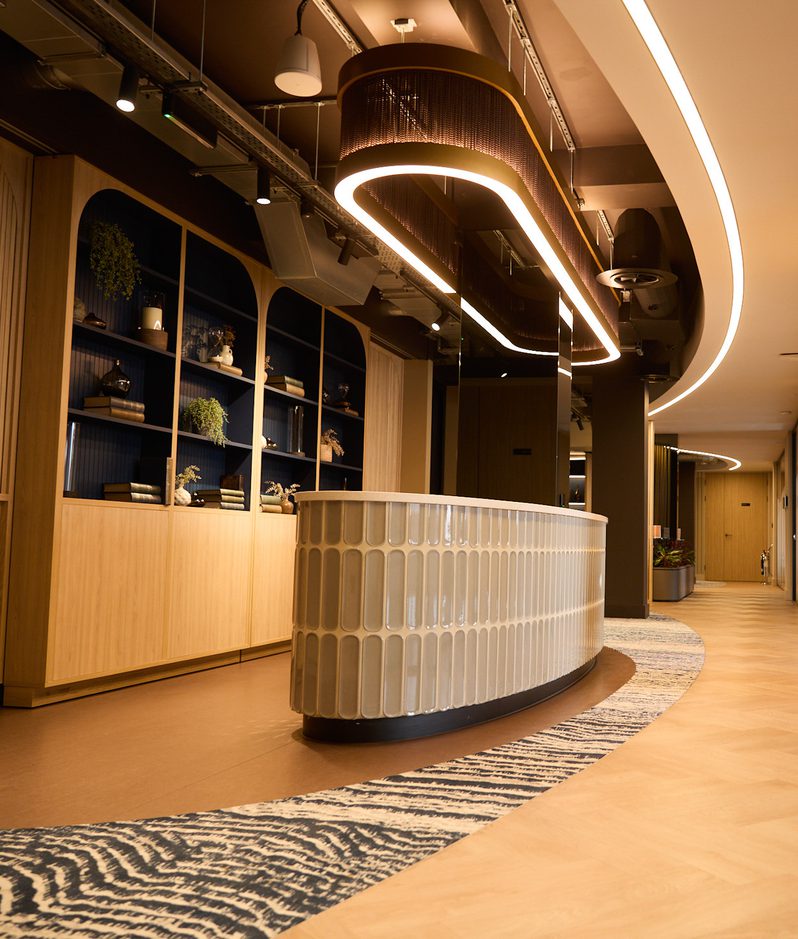 Modern office reception with a curved white textured counter, shelving, ring lighting, zebra-pattern rug, and a long hallway with frosted glass panels.