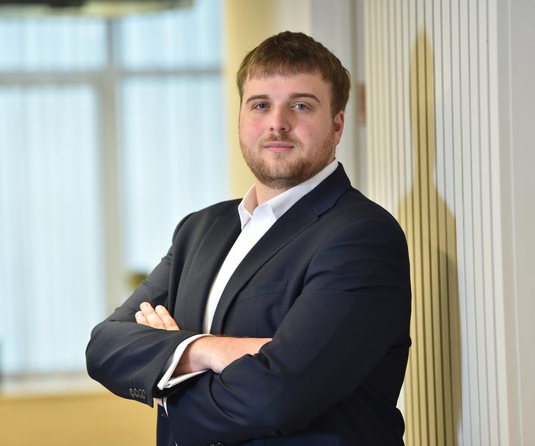 Young man with brown hair and beard, wearing a dark suit and white shirt, standing with arms crossed in an office environment with windows and vertical blinds.