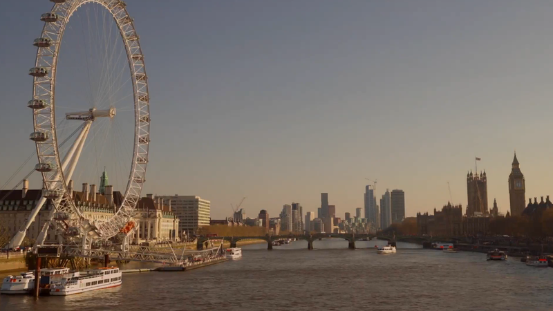 A view of the London Eye ferris wheel on the left, with the Thames River holding several boats, and the city skyline including Big Ben and the Houses of Parliament in the distance.