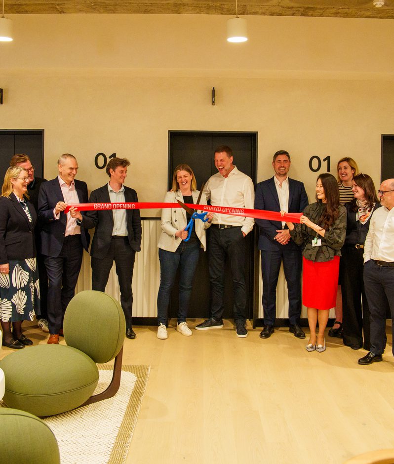 Group of professionals in smart attire gathers for a grand opening ribbon-cutting, as a red ribbon is cut in front of a modern reception area with seating and beige walls.