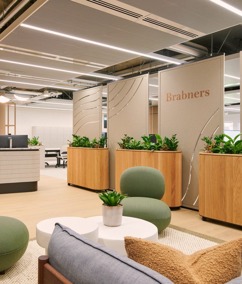 Modern office lobby with a curved tiled reception desk, wooden planters, green lounge chairs, potted plants, and beige divider panels bearing Brabners
