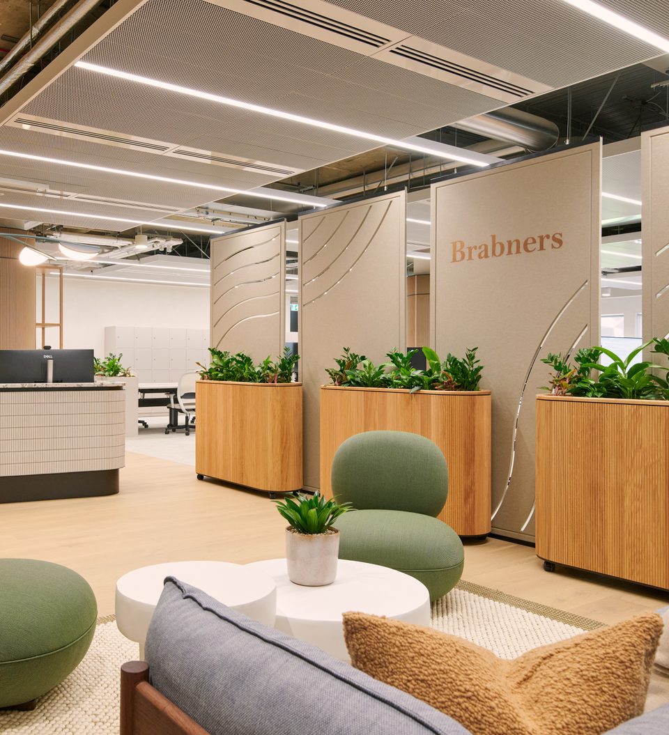 Modern office lobby with a curved tiled reception desk, wooden planters, green lounge chairs, potted plants, and beige divider panels bearing Brabners