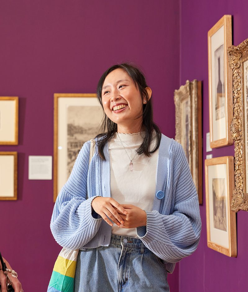 Two people chat in an art gallery with a purple wall, smiling at framed artworks; one in a black sleeveless top and beige trousers, the other in a light blue cardigan and white top.