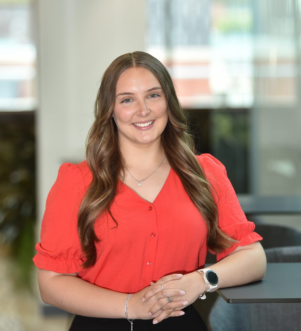 A woman with long wavy brown hair, wearing a red blouse, is smiling and standing indoors near a table, with a modern office background behind her.