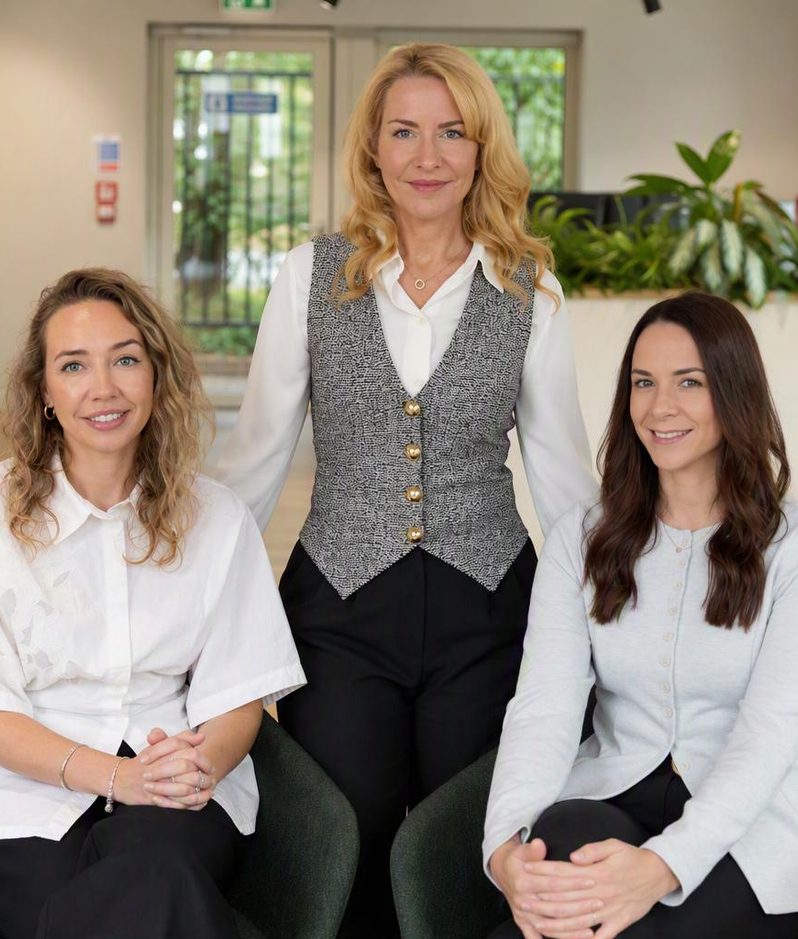 Three people smile in a bright office lobby with plants; a person in a grey patterned waistcoat over a white blouse stands between two seated colleagues.
