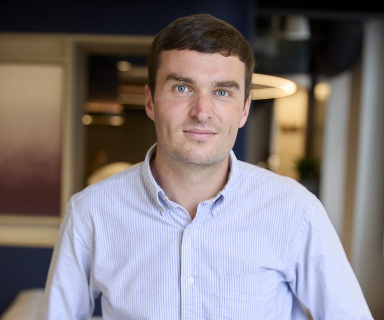 Person with short dark hair and blue eyes, wearing a light blue striped shirt and dark trousers with a brown belt, standing in a modern office setting.