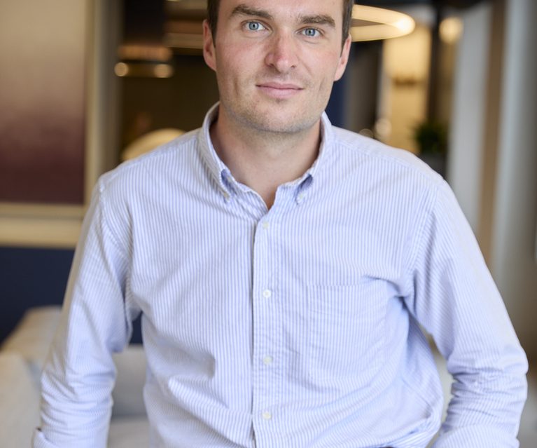 Person with short dark hair and blue eyes, wearing a light blue striped shirt and dark trousers with a brown belt, standing in a modern office setting.
