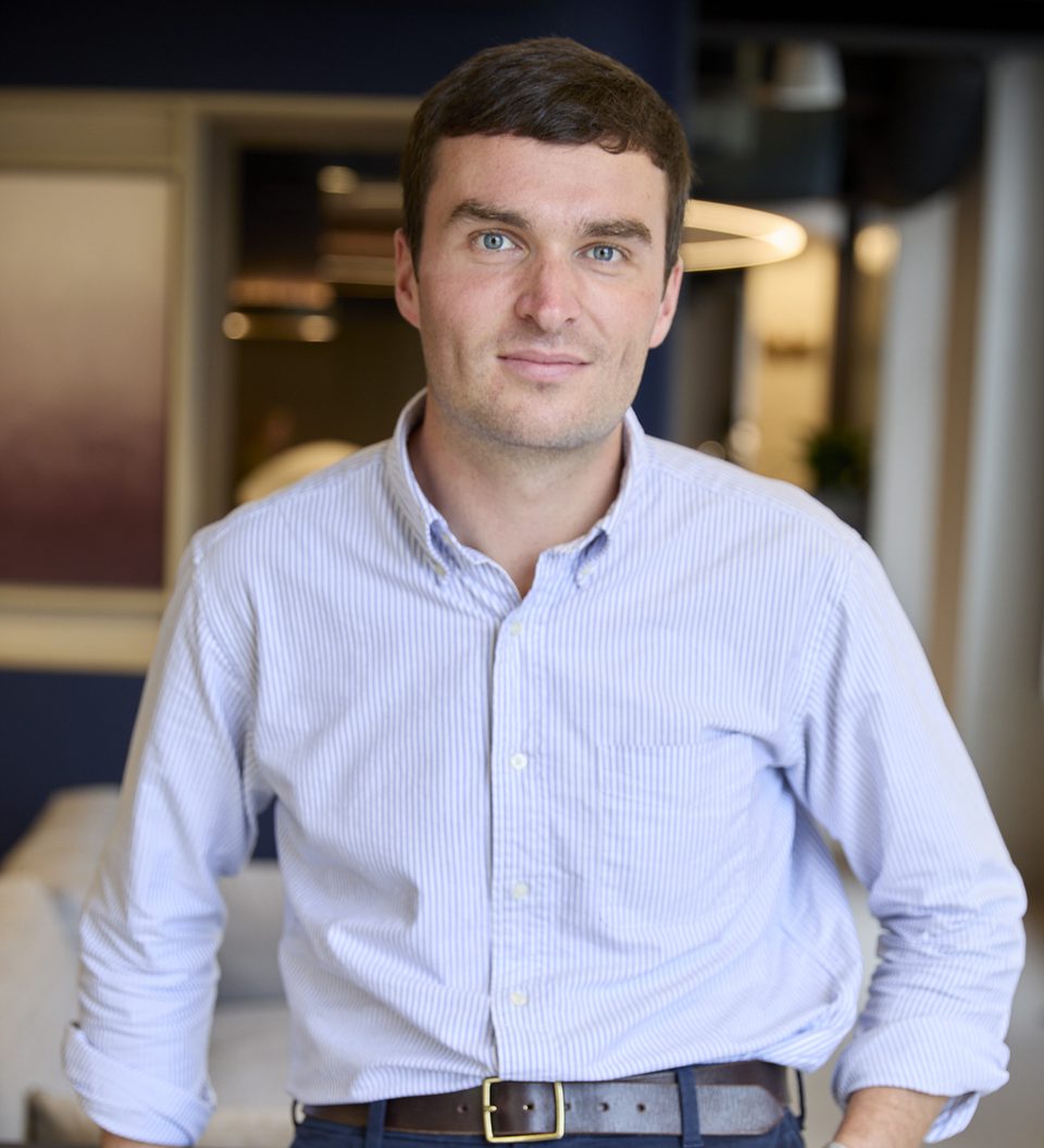 Person with short dark hair and blue eyes, wearing a light blue striped shirt and dark trousers with a brown belt, standing in a modern office setting.