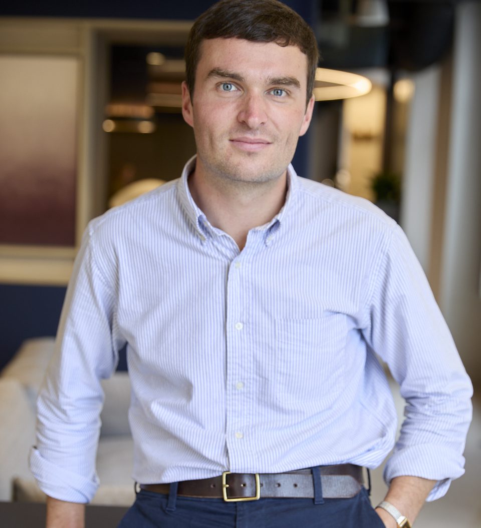 Person with short dark hair and blue eyes, wearing a light blue striped shirt and dark trousers with a brown belt, standing in a modern office setting.