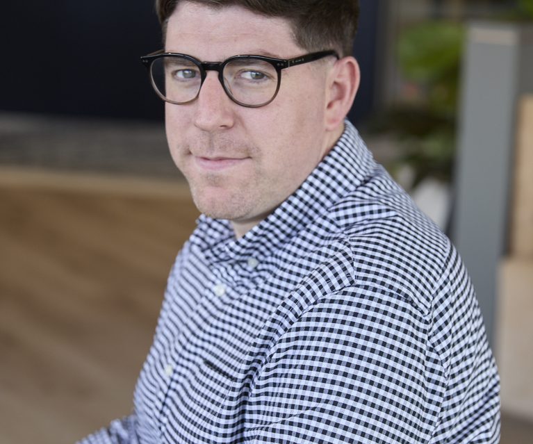 Portrait of a person with short dark hair and round black glasses, wearing a blue and white gingham shirt, looking at the camera in a modern indoor space.