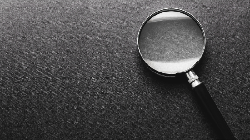 Grayscale close-up of a magnifying glass with a black handle resting on a textured dark surface; the round lens catches light along its rim.