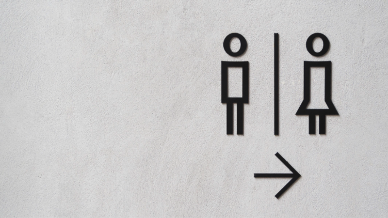 Two simple stick-figure icons separated by a vertical divider with a right-pointing arrow, set on a light textured wall, resembling a toilet sign.
