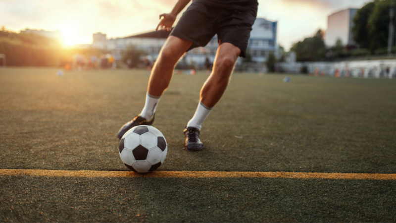 A person is preparing to kick a soccer ball on an outdoor field at sunset, with buildings and trees in the background.