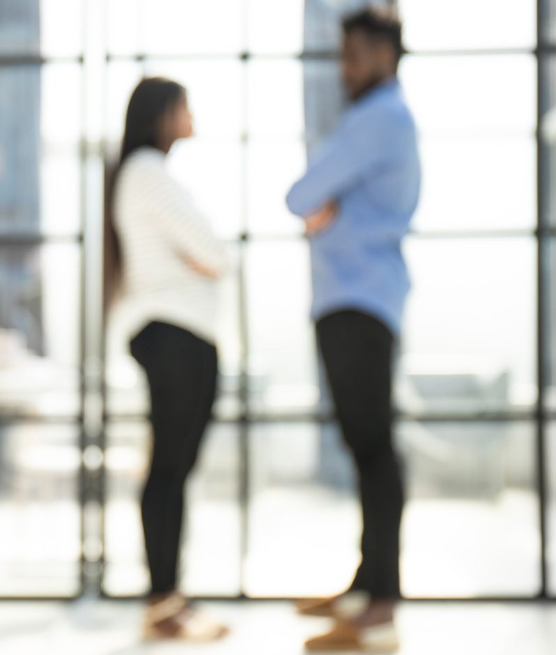 Two people standing and talking in front of a large grid window in a modern office building, with bright natural light and a blurred cityscape in the background.