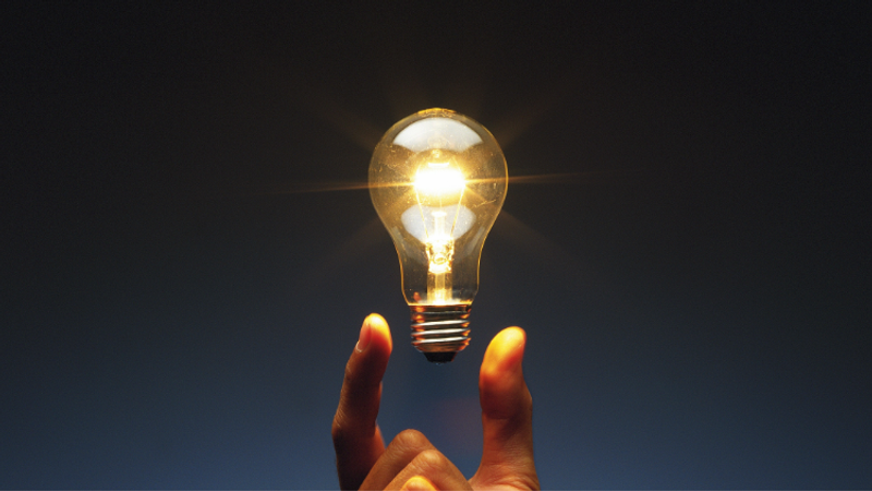 A hand holds a glowing incandescent light bulb, with visible filament, against a dark blue gradient background.