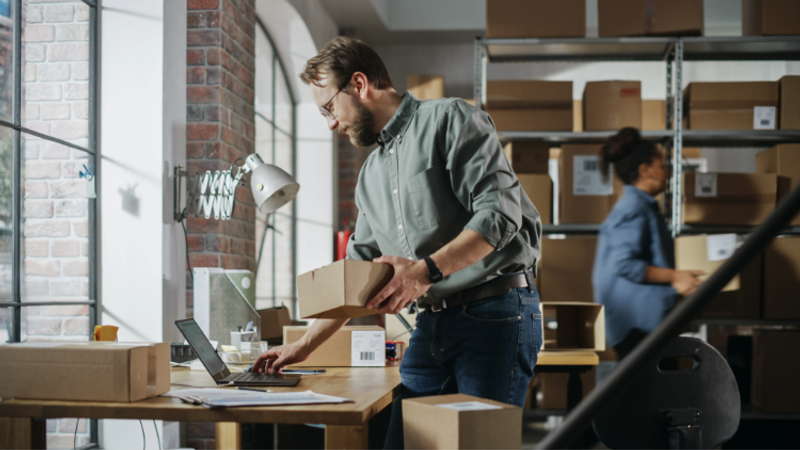 Person with glasses and beard packs a box at a wooden desk in a warehouse, using a laptop; shelves with boxes and another worker in the background.