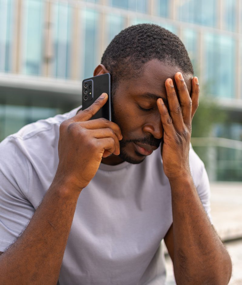 A man sitting outdoors in front of a modern glass building, holding his forehead with one hand and talking on a smartphone with the other.