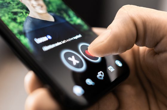 Close-up of a finger pressing the heart icon on a dating app screen; circular X and heart buttons are visible, with a blurred profile photo in the background.