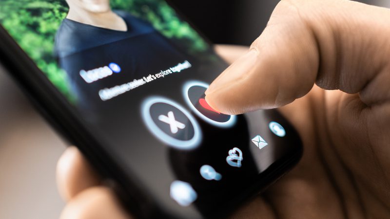 Close-up of a finger pressing the heart icon on a dating app screen; circular X and heart buttons are visible, with a blurred profile photo in the background.