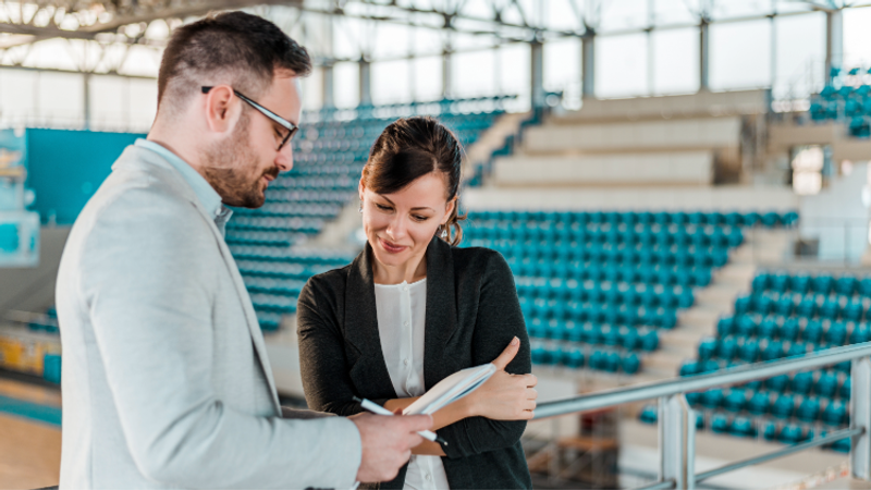 Two professionals review documents together in an indoor arena with teal seating and natural daylight from large windows, standing by a railing as they discuss a notebook.