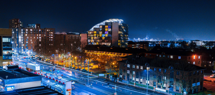 Night cityscape featuring a curved glass-fronted building, busy illuminated streets, and modern brick and glass structures along a wide avenue under a deep blue sky.