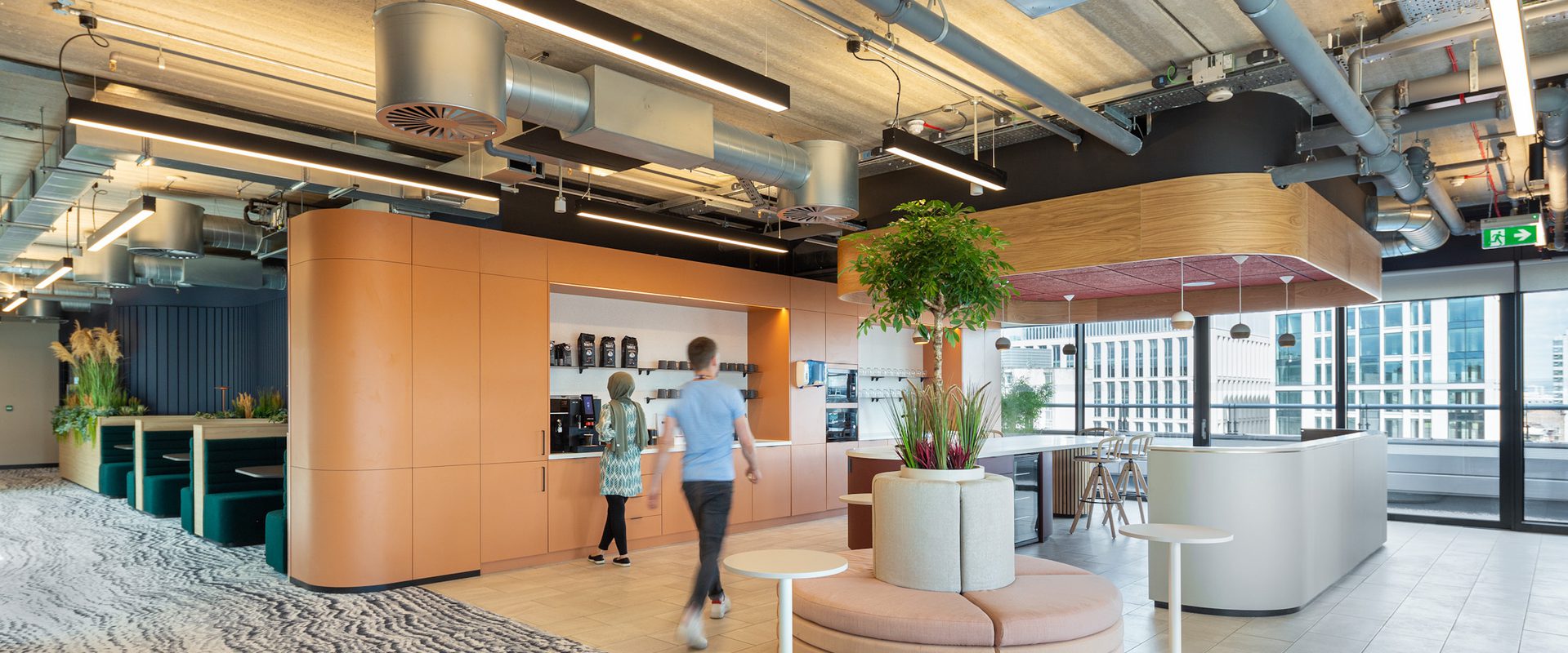A modern interior space with a coffee station, seating area, and large windows showing a city view; ceiling features exposed pipes and ventilation systems.