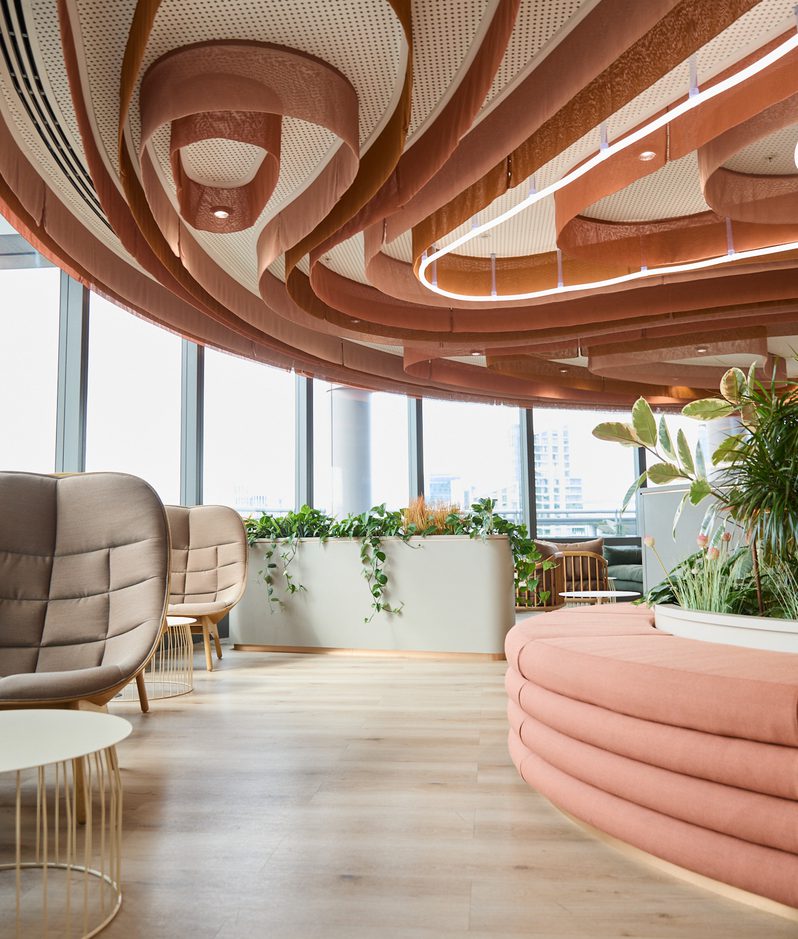 Interior seating area with curved pink upholstered bench, beige lounge chairs, brown fabric spirals from the ceiling, and potted plants by large windows.