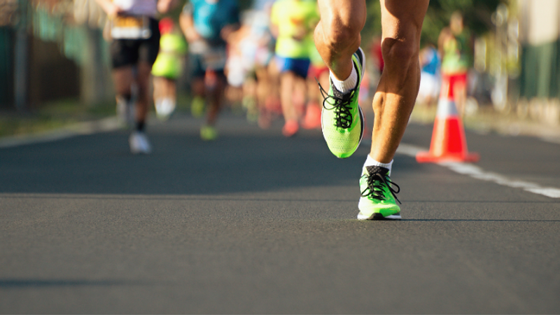 Close-up of a runner wearing bright neon-green shoes sprinting on a city street, with blurred runners in the background and an orange traffic cone on the right.